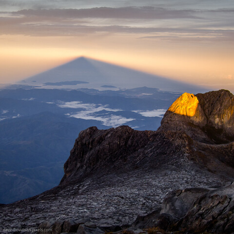 A colour photograph of sunrise from the summit of Mount Kinabalu, the highest peak in Southeast Asia, located on the island of Borneo in the Malaysian state of Sabah, looking down the mountain to the land below. The early morning sun's rays cast a triangular shadow from the mountain onto atmospheric mist.