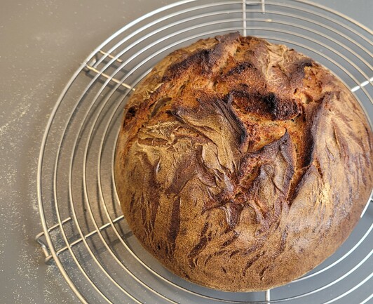 Foto: Auf einem Kuchengitter liegt ein rustikaler, runder Laib Brot mit in der Mitte kräftig aufgerissener, leicht bemehlter Kruste. Die Kanten am Ausbund sind dunkler als der Rest und sehen schön knusprig aus. 