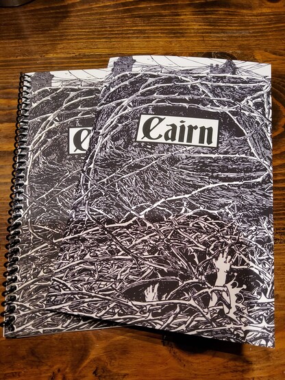 Picture of the covers of the spiral-bound and staple-bound copies of the Cairn role playing game by Yohai Gal. The cover features the Cairn title in a Gothic font on top of a black and white illustration of a thick bramble with a crown a hung within it and a hand reaching out of it in the lower left corner. The impression of a castle or keep can be seen in the distance beyond the brambles. 