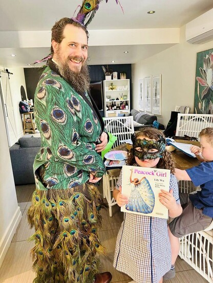 Photo of the author dressed in a three piece Peacock suit, peacock wedding train with bustle and peacock Mohawk. Standing next to them is a young girl in a school uniform wearing a peacock masquerade mask and holding a copy of the book Peacock Girl. In the background is a young boy working at the kitchen table in the background.