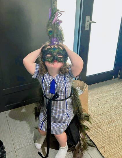 Young girl in school uniform wearing a peacock Mohawk, peacock masquerade mask and peacock wedding train with bustle. She is crouching down slightly, face on with her hands held up on either side of her head.