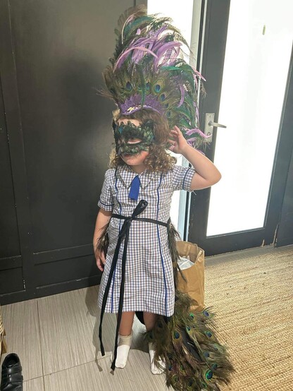 Young girl in school uniform wearing a peacock Mohawk, peacock masquerade mask and peacock wedding train with bustle. They are posing partially side on with right arm lifted up and touching the back of her head.