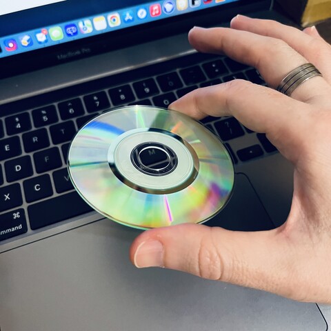 A Mini CD being held between the thumb and index finger of my right hand.  A ring is visible on my middle finger and the disc’s pale green reflective data side is facing up with a rainbow prismatic pattern radiating from its centre.

I’m the background under and behind the disc and hand is the keyboard, track pad and screen of my laptop.