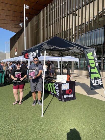 Two people in Headcount shirts holding Headcount clipboards in front of the Headcount tent outside the Moody Center venue in Austin, looking forward to getting people registered to vote and then seeing Foreigner live in concert!!