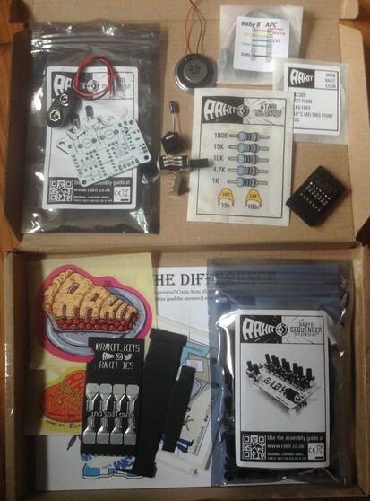 Photo of two electronics kits (a Baby8 and Atari Punk Console) from RaKit.  One has been opened showing neat paper envelopes with cool graphics showing the components inside.  The only sits alongside colourful stickers and merch for RaKit.  Everything is sat in a flat cardboard box for postage.