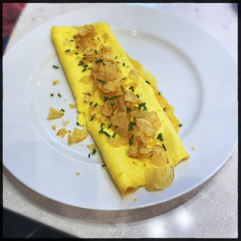 An omelette made in the French style, topped with crumbled potato chips and chives in the middle of a white plate. 