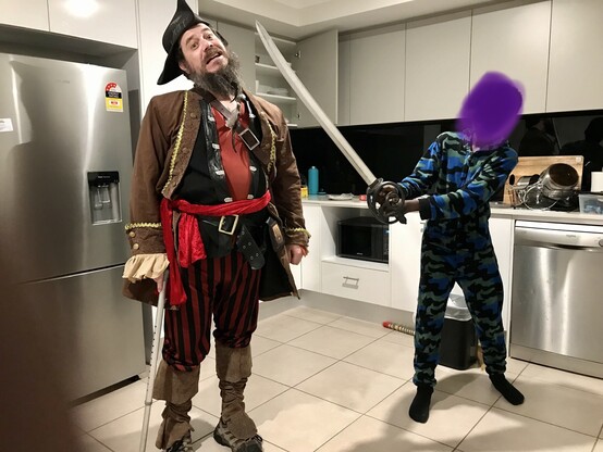 Author dressed as a pirate with tricorner hat, feathers in beard, multiple layer ruffled shirt and jacket and multiple belts. On the right is a young person with outstretched arms holding a long pirate sword to the neck of the pirate.