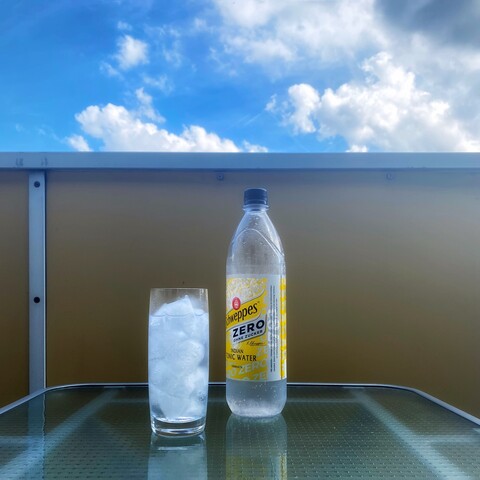 A highball glas with ice and tonic and a bottle of Schweppes Tonic Water Zero on a table, blue sky above.