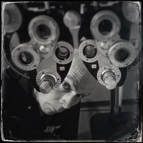 Black and white photo of a man peeking below a phoropter (that multi-lensed eye exam device).