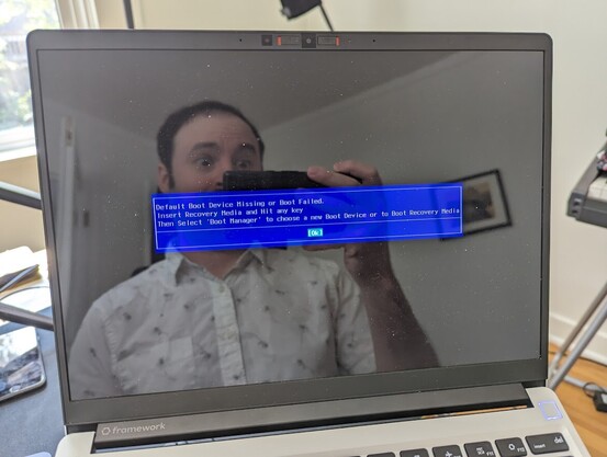 A laptop with a blue BIOS screen reading:  Default Boot Device Missing or Boot Failed  The photographer is making a silly surprised face in the screen reflection.