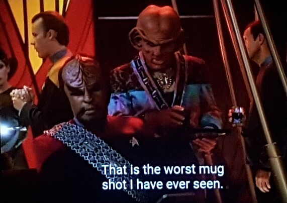 Quark and Worf are in a busy bar on DS9. Worf, in red uniform ,is sitting and looking irritated as per usual. Quark, wearing a jacket made from the leftover fabric scraps bin, is standing over him holding a drink tray and looking at something in Worf's lap. There are random bar folk barfolking in the background. Closed caption reads, "That is the worst mugshot I have ever seen."