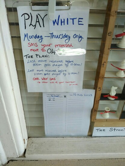 Notice to passers by reads:

Play White.
Mon-Thurs Only.
SMS your proposed move to 04 [rest of number scribbled out]

The Plan
Last move received before 8am gets played by 10am.
Last move received before 2pm gets played by 4pm.

One way SMS
No other use of your number.
Or mine.

Below the notice is an A4 sheet showing the moves that have been made in long algebraic format. (Long is more accessible to beginners)