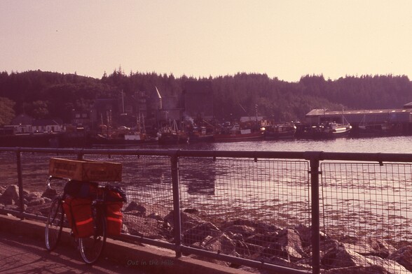 Bicycle laden with panniers and a wooden fish box leaning against railings, with a rocky shore and the sea behind. At the back some buildings and a few fishing boats moored. Tree lined slopes beyond.