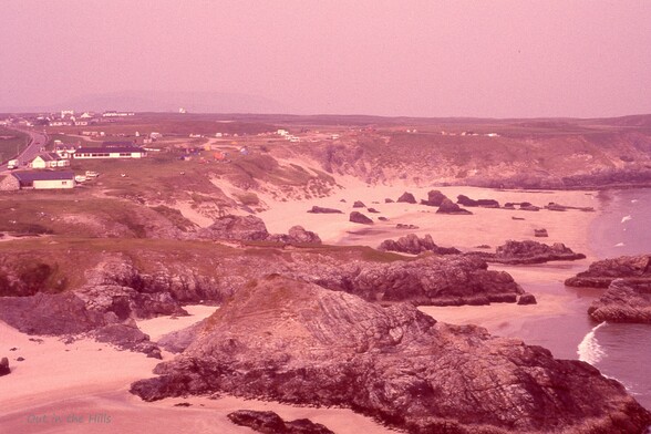 A hazy view of a coastline. Sandy beach with rocky outcrops and dunes. A spread out village behind the beach.