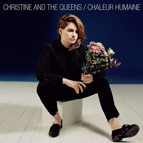 Album cover de l’album Chaleur Humaine de Christine and the Queens. Assis sur un cube blanc, habillé de noir avec un bouquet de fleurs dans les mains. Le sol est un parquet blanc et le mur est bleu foncé. 