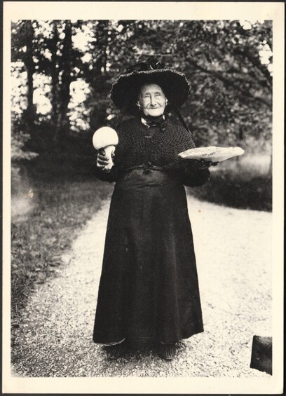 A black and white postcard of an elderly woman on a road, dressed all in black and holding two mushrooms, one round and bulbous and one large and saucer-shaped.
