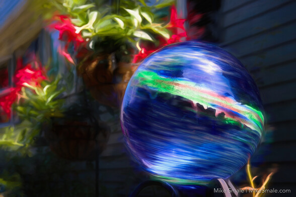 A closeup of a blue gazing ball with a green stripe through its centre against a green house and red mandevilla in hanging pots.