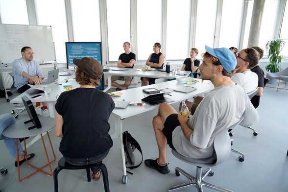 A group of 10 people in informal clothes sitting around a table arrangement in a modern light-flooded office – with a whiteboard and a poster of the Service Standard behind them
