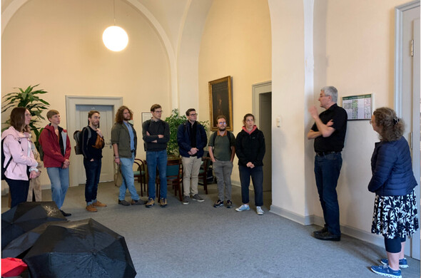 Nine young people standing in a big hall and listening to a grey-haired man, in the front open umbrellas laying on the ground.