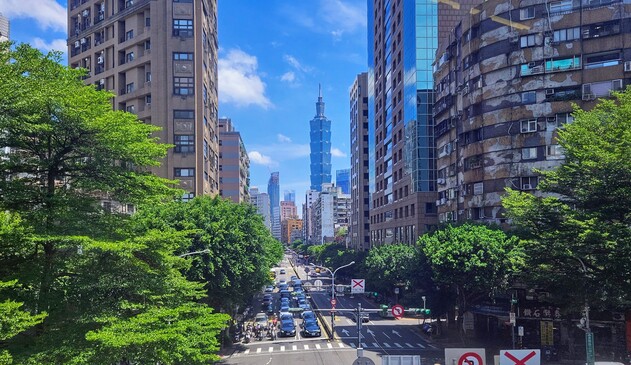 Blick durch eine Allee aus Baeumen und Wolkenkratzern hin zum Taipei 101, der im Hintergrund steht. Strahlend blauer Himmel