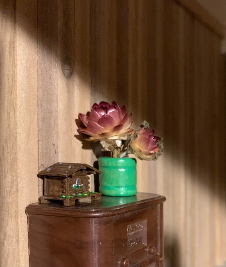 A tiny hinged chalet on top of a miniature radio, along with a small vase of flowers. They are in a Lundy-sized dollhouse.