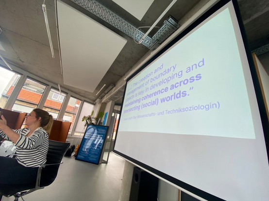 A woman in a large office space with a large projected quote projected behind her: “The creation and management of boundary objects is key in developing and maintaining coherence across intersecting (social) worlds” Susan Leigh Star