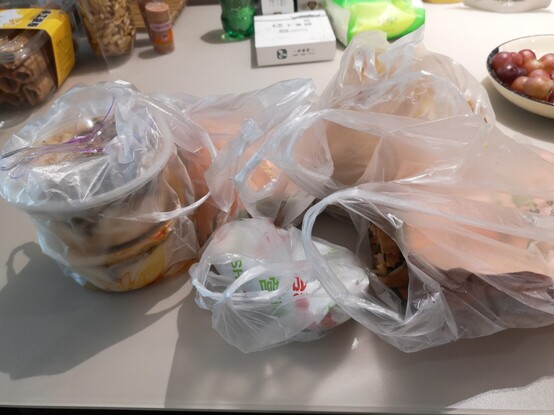 Foto von Frühstück, welches bei Straßenhändlern gekauft wurde.
Zu sehen sind mehrere Plastiktüten,in denen sich kleinere Plastikbecher mit Suppen oder in Papier eingewickelte Speisen befinden.
Die Speisen stehen auf einem Tisch.