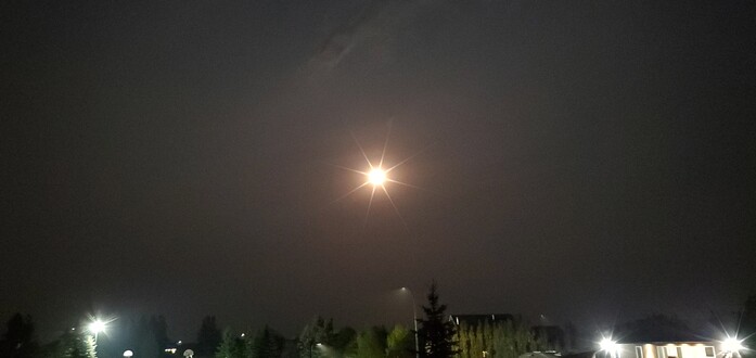 The Moon radiates in the night sky with residential buildings and lights at the bottom of the picture
