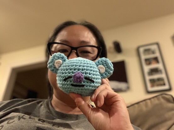 A crocheted blue koala head, about 5 inches wide, with a purple nose and a sleepy, slightly smiling face. I’m holding it in front of my face. 