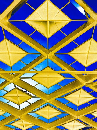 A picture of the metal and glass roof above the tram platforms at the central railway station in The Hague. There are square metal pieces set in large square holes, held in place by narrow ties, leading to a strong geometrical pattern of diamonds from this angle. The image has been solarised to turn the originally dark metal squares bright yellow-gold, while the sky seen through the glass sections is a deep blue.