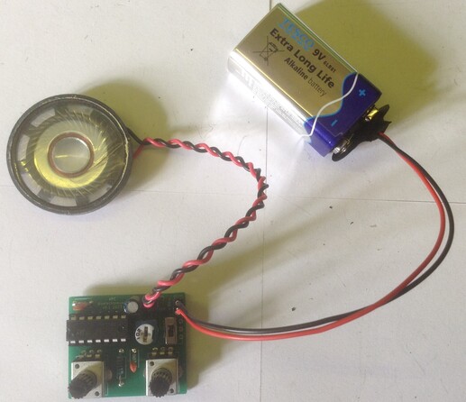 Photo of a small custom PCB (bottom) with a 14-pin chip and two potentiometers connected to a small headphone loudspeaker and a 9V battery.