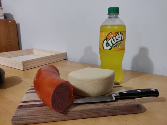 Kielbasa sausage and friulano cheese on a cutting board with a knife. A bottle of pineapple Crush in the background.