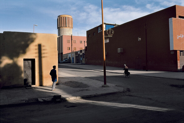 A shot of a street in strong sunlight. In the centre of the frame a person is standing dead still on the pavement.