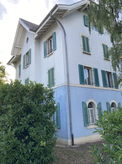 The same place as the following picture, focussing on the house. The house is now painted in light colours with green shutters, some of the decorative gabling has been removed. Tall trees are growing around it, partially obscuring the facade. But the shape remains and it is noticeably the same distinctly-shaped building.
