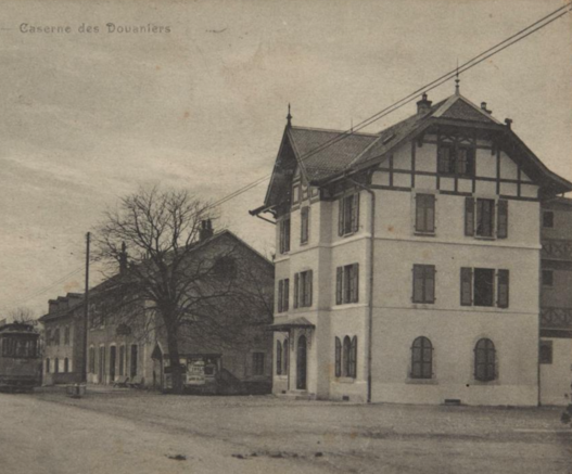 An archive sepia picture of the Caserne des Douaniers in Perly, near Geneva, dating from before 1914. The road is broad with old houses two-story houses. The Caserne was built to house border guards, in a distinct three-storey style with decorative gabling applied below the roof.