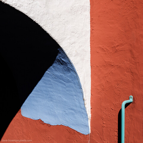 A semi-abstract photograph of a section of red, white and blue wall with a light blue metal handrail attached to it in the village of Portmeirion in Wales, UK. The wall is lit by bright summer sunlight.