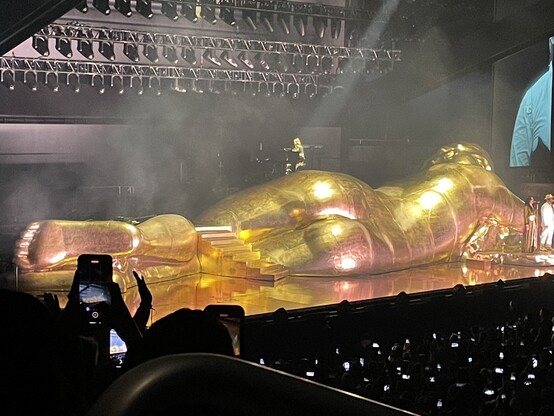A giant nude metallic cherubic figure stretches all the way across the stage. We see his backside and prominent buttocks.