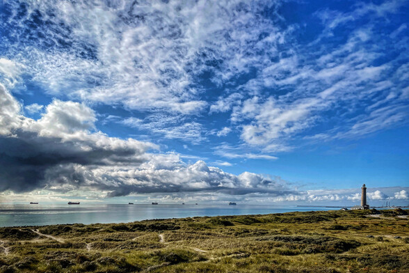 Dunes, the calm Baltjc Sea, some ships on the horizon and a dramatic blue sky with some clouds in Skagen, Denmark. On the right, Skagen‘s lighthouse is visible in the distance. 