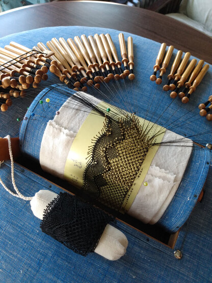 A bobbin lace roller pillow, shot from the back. It's an oval sort of platform, with a cylinder in the middle that turns as you work your pattern around. A little holder in the back winds the completed yardage. 

It is a black silk lace, about 2 inches wide. The fancy edge has kind of a wavy look with little picots off the side. In each wave trough a small diamond motif forms. The rest of the lace is the ground stitch, just like the background that lets the motifs show up. 

The completed part has pins removed. The recent work still has brass pins at each stitch location to hold them until you get further along.

Towards the upper part of the photo, the 50 or so bobbins wound with the unused thread are waiting. 