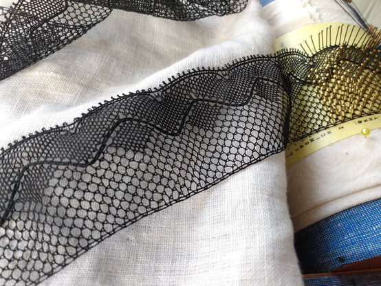 An opened up piece of my completed black silk lace, unwound  to see the detail. 

About 2 inches wide, it has one side that has a wavy motif with a heavy border. The outermost edge has little picot twisted bits.  In the trough of each wave is a little diamond shape stitch area. 

The other edge is called the sewing edge, and you'd put something like this on your black shawl or cape edge using that sewing edge. It just has an extra close thread to make a border at the edge. 

It's just laid out on a white linen fabric for contrast. It's still attached to the working section over on the right so it's hard to photograph well. The working section has brass pins in the yellow pattern paper where the stitches get made. It's on a roll that I can turn as I complete a segment. 