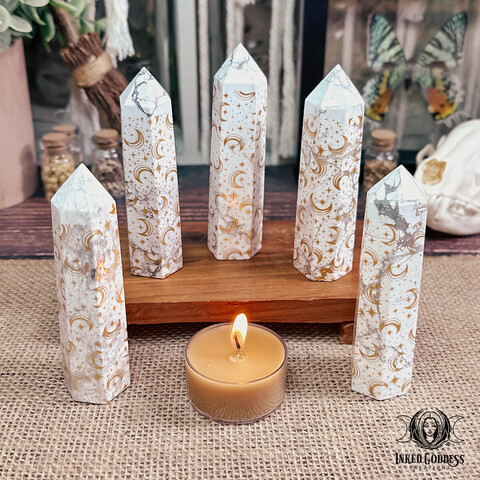 A photo of our "Howlite Moon Tower" product. It features three specimens on a wooden platform, with another two specimens atop a wood table covered in beige burlap. A gold tealight candle is shown for scale. Presented by Inked Goddess Creations.