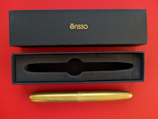 A posted Ensso PIUMA brass fountain pen alongside its simple black cardboard and foam case on a textured red mat.