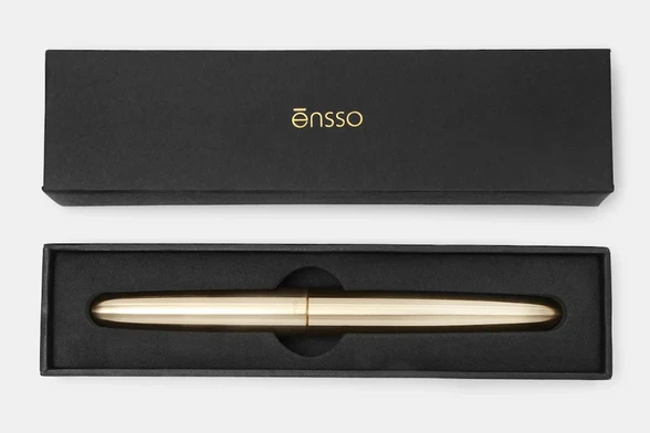 A product photograph of a posted Ensso PIUMA brass fountain pen in its simple black cardboard and foam case.