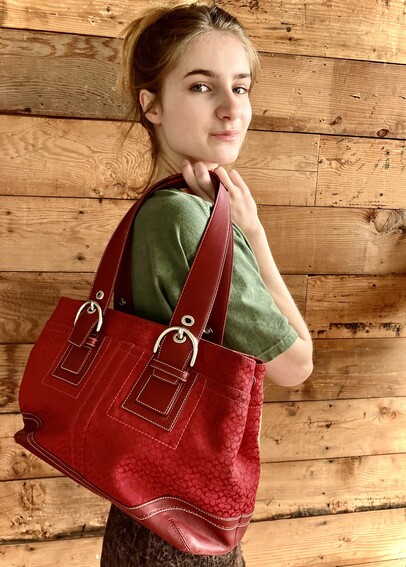 A slender, honey-blonde-haired young woman holds a bright red purse over her shoulder in front of a shiplap wood wall. 