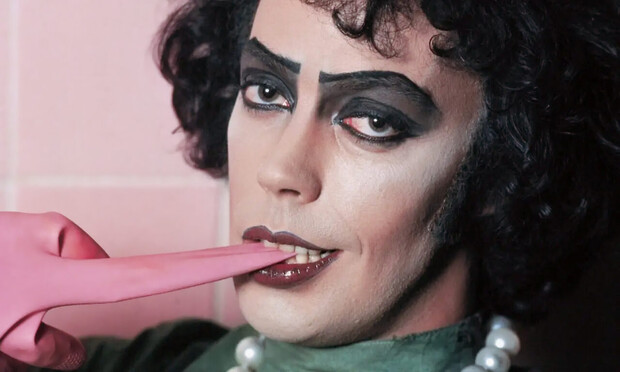 Man with long, curly black hair, black eye shadow and red lipstick looks at the camera and bites the thumb of a pink rubber glove, pulling it off his hand. Tim Curry as Dr. Frank-N-Furter in a still from Jim Sharman’s 1975 movie “The Rocky Horror Picture Show“ based on Richard O’Brien’s musical The Rocky Horror Show.