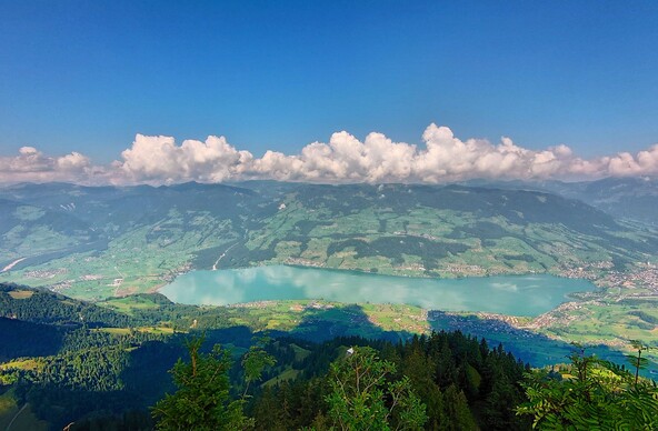 Blick hinab auf den Sarnersee. Dahinter Berge vor denen Wolken hängen. Alles ist grünblau. 