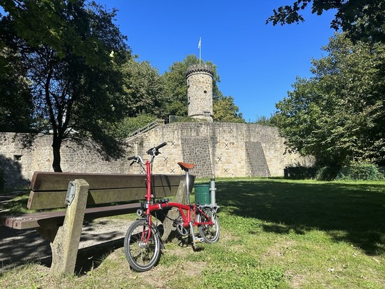 A tower and wall of the castle ruin Tecklenburg. In the front there is  a park bench and  a red folding bike.