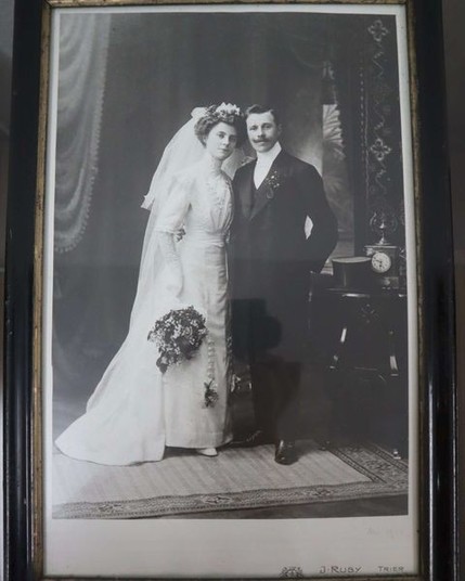 Ein Hochzeitsfoto von 1912, Die Braut trägt das besagte Kleid einen Schleier nach hinten und weise Handschuhe in denen sie einen Blumenstrauss hält. Ihre blonden Haare sind in einer gewellten Hochsteckfrisur drapiert.
Der Bräutigam trägt einen Schwarzen Mantel mit Blumen im Knopfloch mit schwarzer Weste. Seine schwarzen Haare sind kurz geschnitten und zur Seite gekämmt, er trägt einen mittelgrossen Schnurrbart.