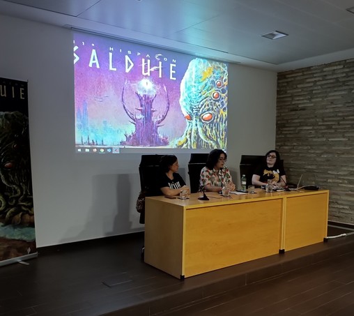 Caryanna, Darkor y Dikana en la mesa de la sala de conferencias con una pantalla detrás donde se proyecta el cartel de este año de la Hispacon