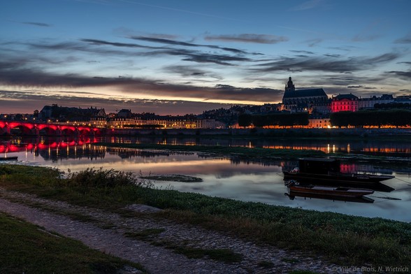 Photo depuis le port de la Creusille au coucher de soleil : le pont Jacques-Gabriel et l’Hôtel de Ville sont illuminés en rouge et se reflètent dans la Loire.
